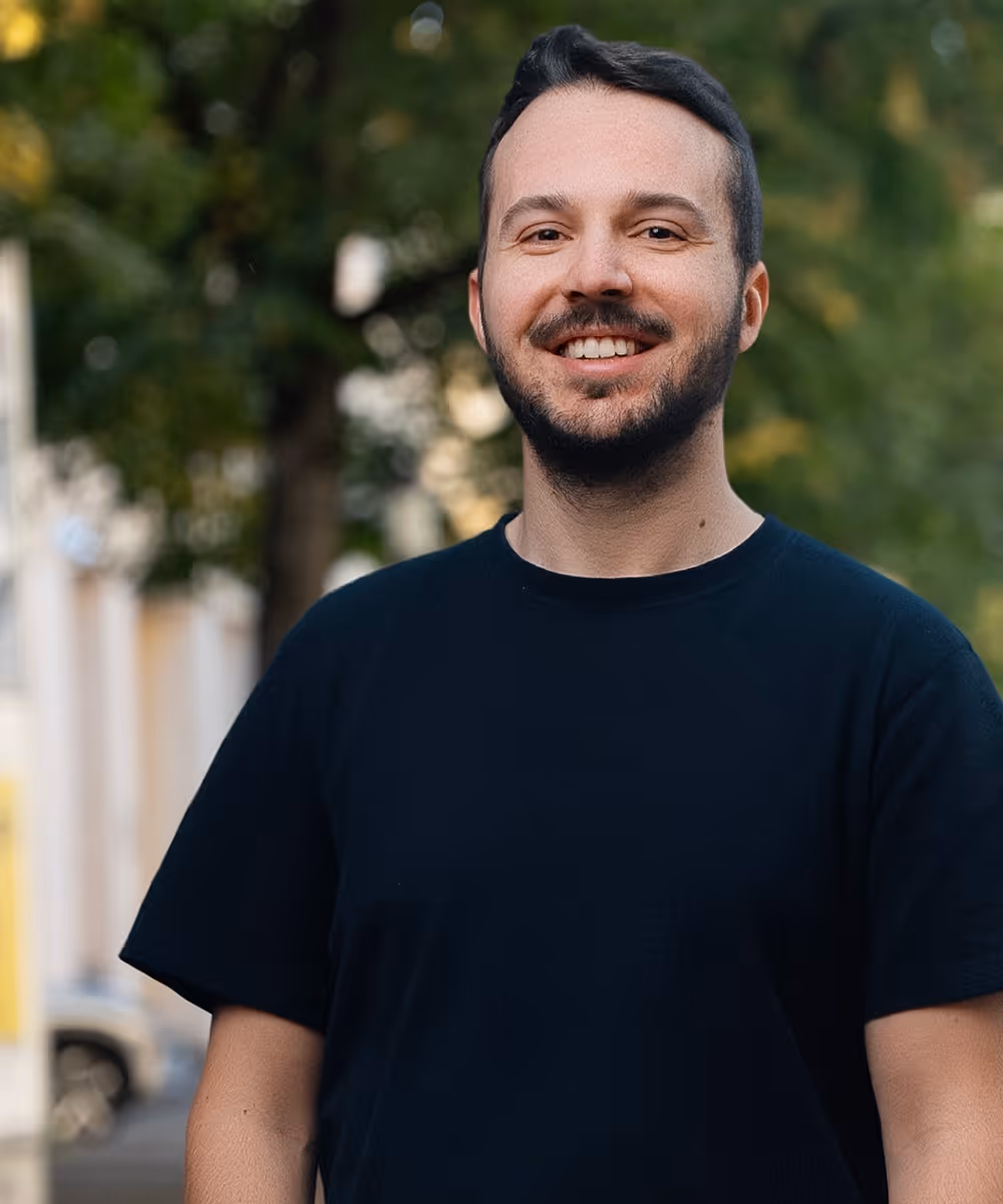 Lächelnder Mann mit kurzen dunklen Haaren und Bart, trägt ein schwarzes T-Shirt, im Freien vor grünen Bäumen.
