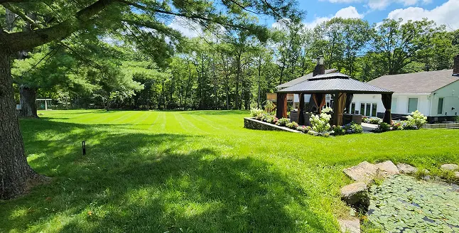 Well-maintained green lawn with a shaded gazebo near a white house, surrounded by trees and a pond with lily pads.