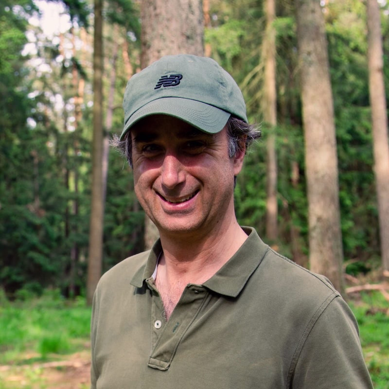 Mann mit grüner New Balance Kappe und Poloshirt lächelt in einem Wald.