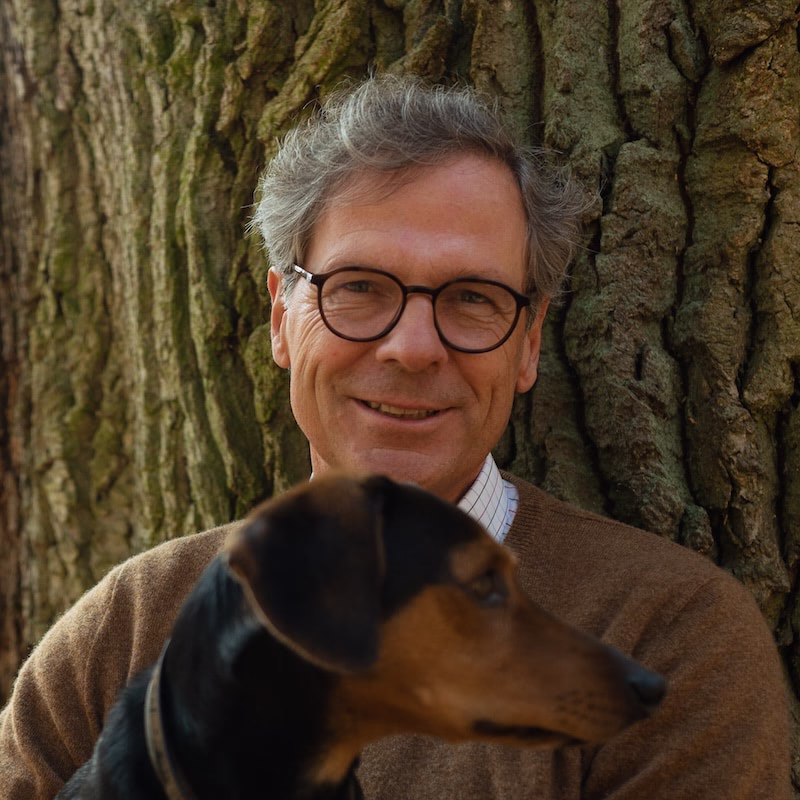 Mann mit Brille und grauem Haar lehnt an einem Baumstamm, im Vordergrund ein braun-schwarzer Hund.