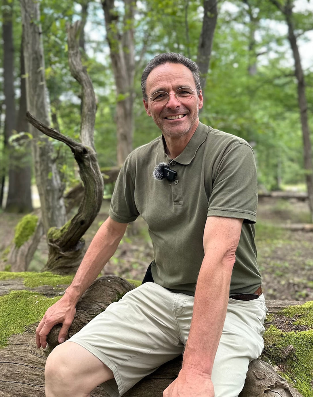 Mann mit Brille in grüner Polohemd und kurzen hellen Hosen sitzt lächelnd auf einem moosbedeckten Baumstamm im Wald.