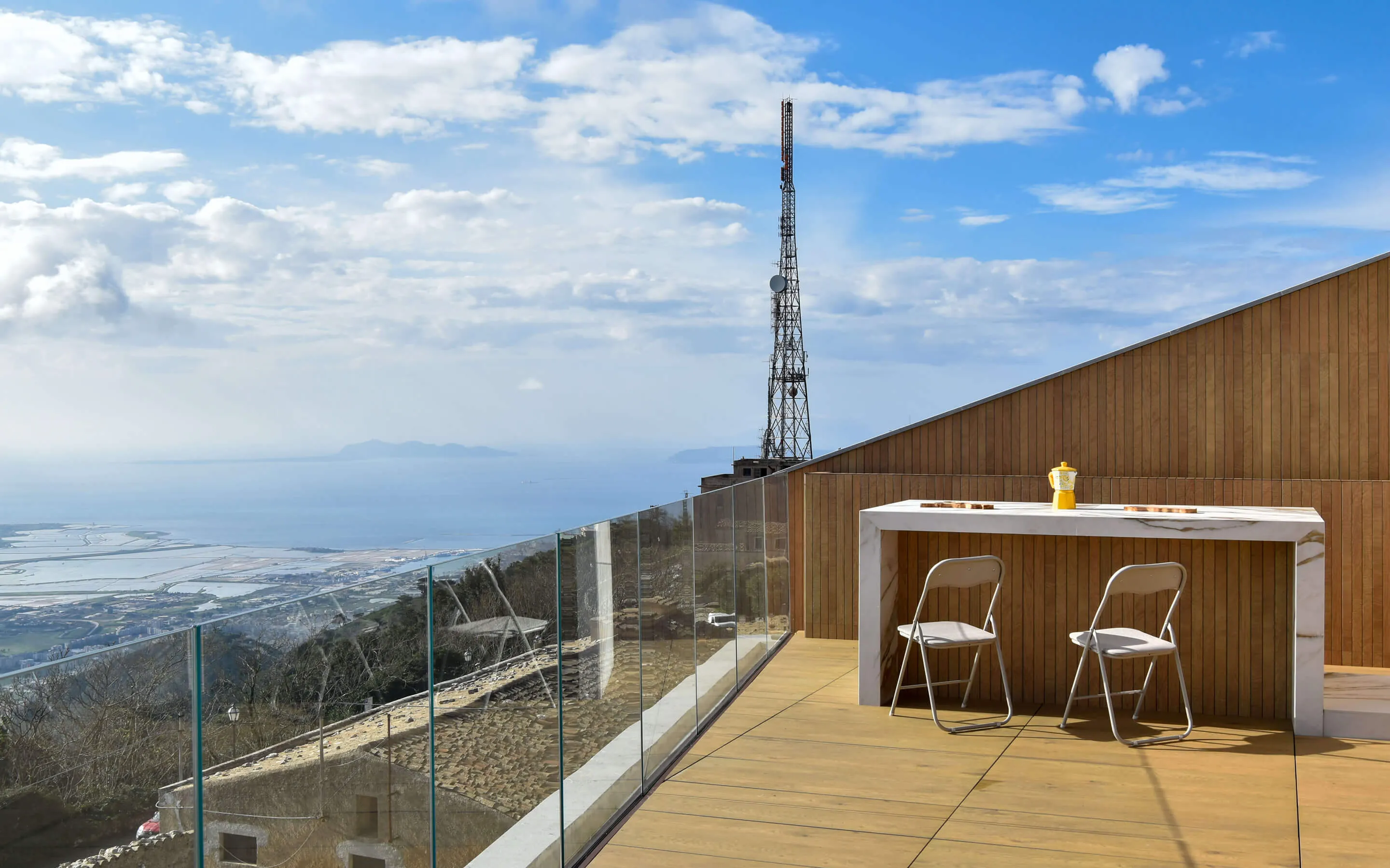 Outdoor terrace with glass railing, two chairs, a table, and a distant ocean view under a blue sky.