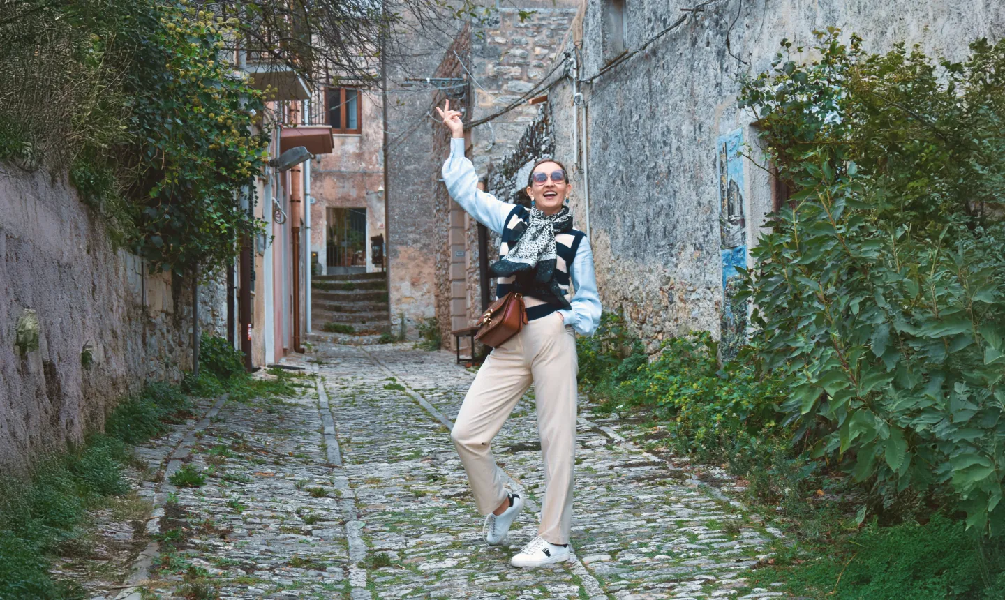 Woman posing happily on a cobblestone street lined with old stone buildings and greenery.