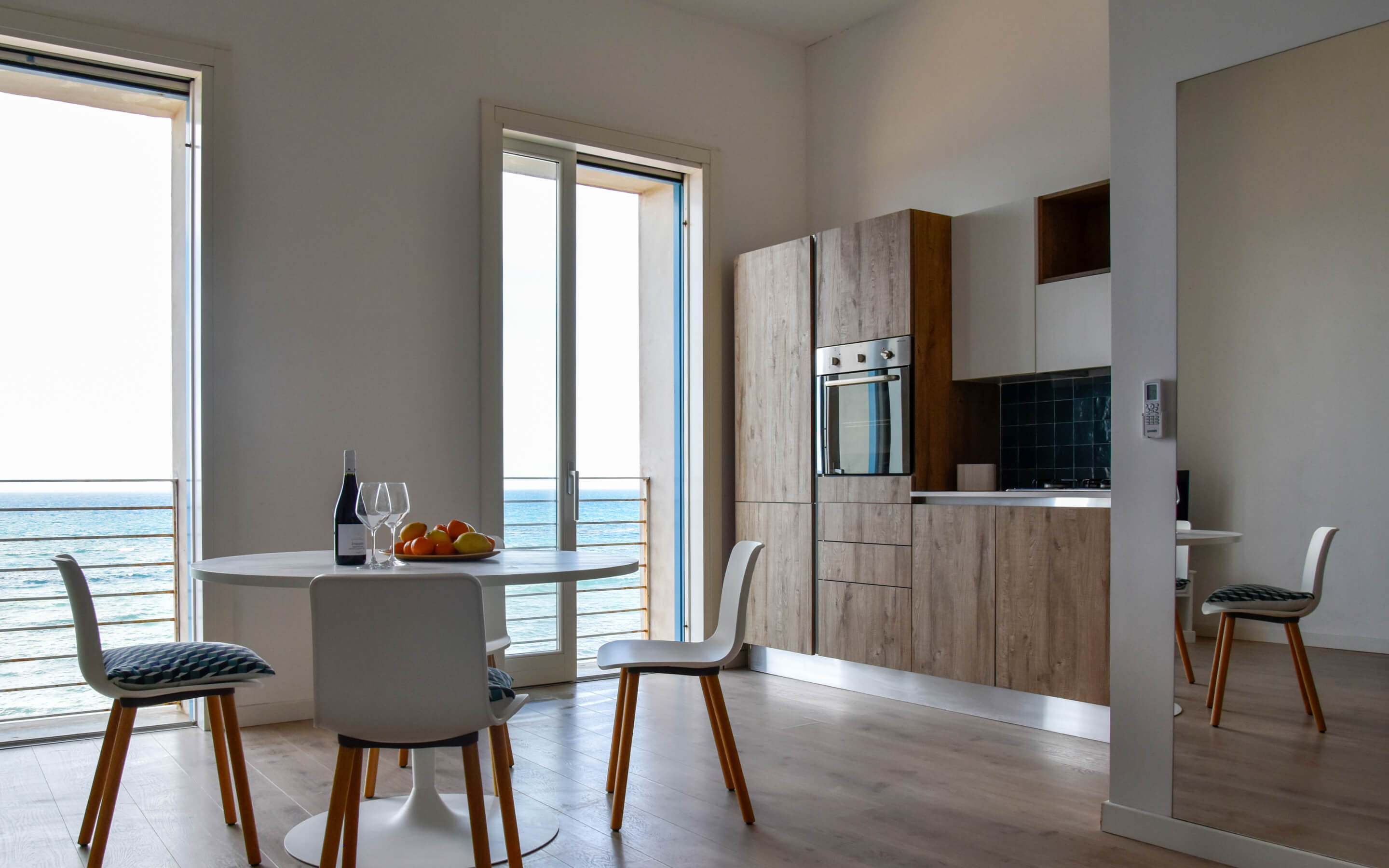 Modern kitchen with wooden cabinets, round table with chairs overlooking ocean through glass doors.