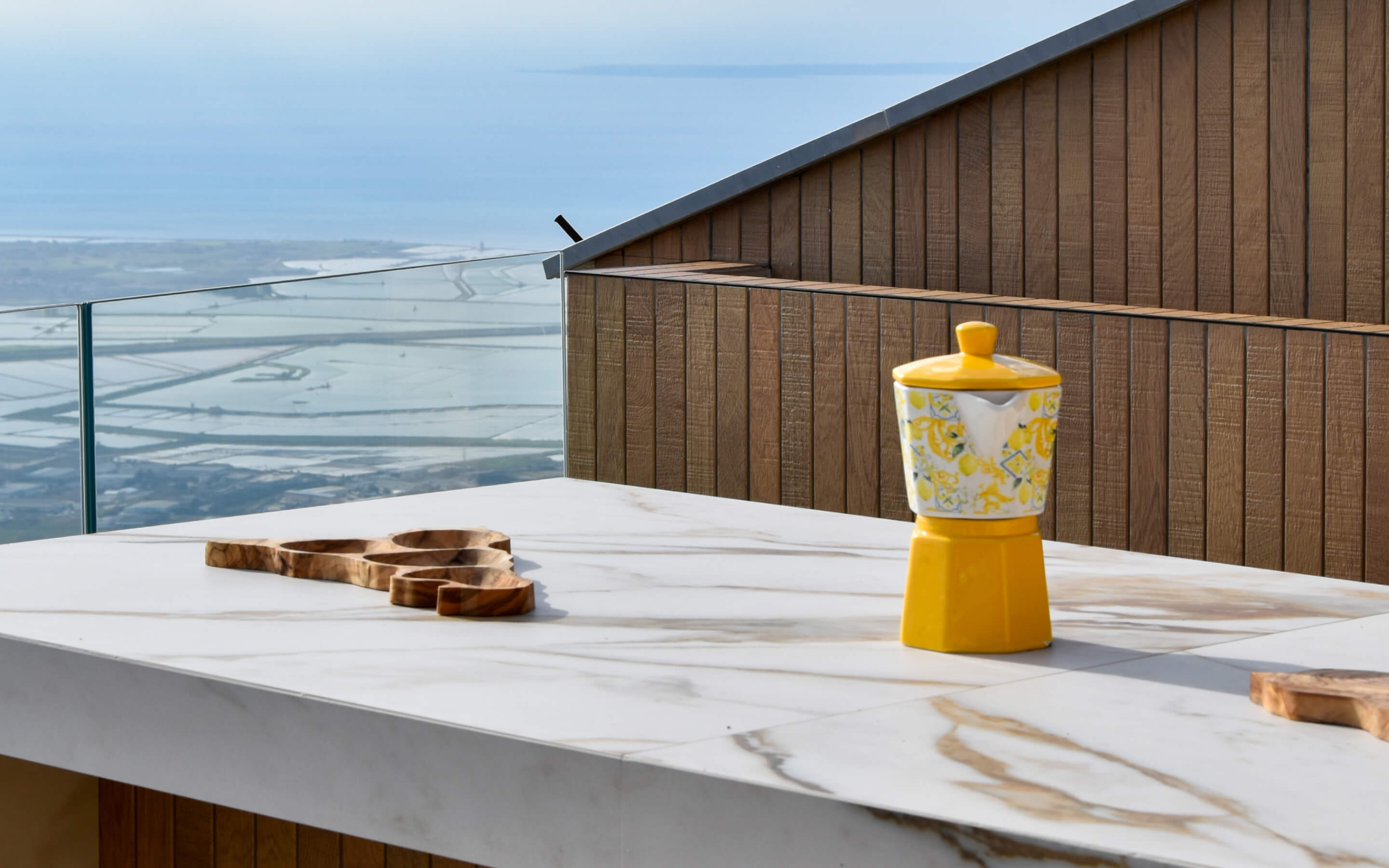 Yellow patterned stovetop coffee maker on a marble countertop against a wood-paneled wall and glass railing with sea view.