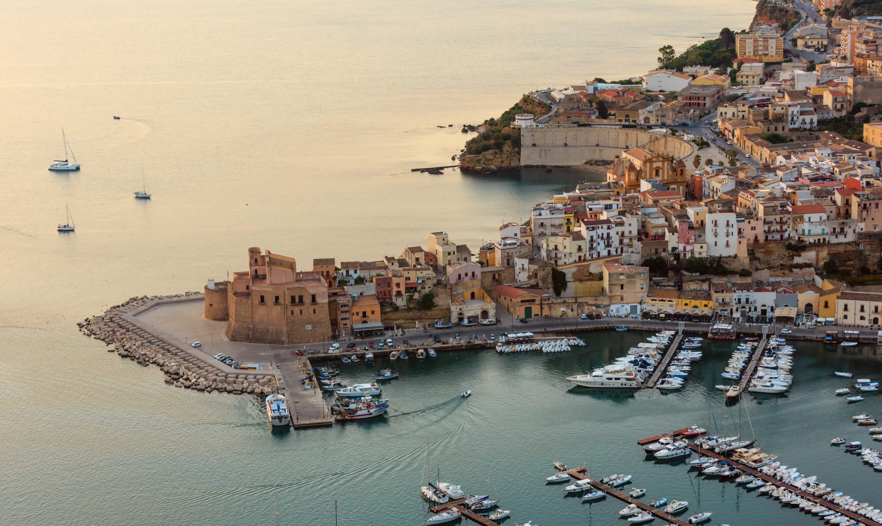 Aerial view of a coastal town with a marina, boats, historic buildings, and calm sea at sunset.
