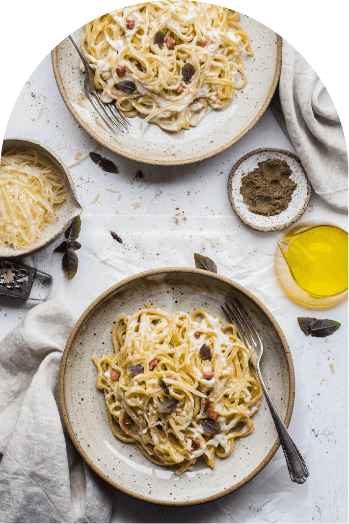 Two plates of creamy pasta with herbs, grated cheese, and a small dish of pepper and olive oil.