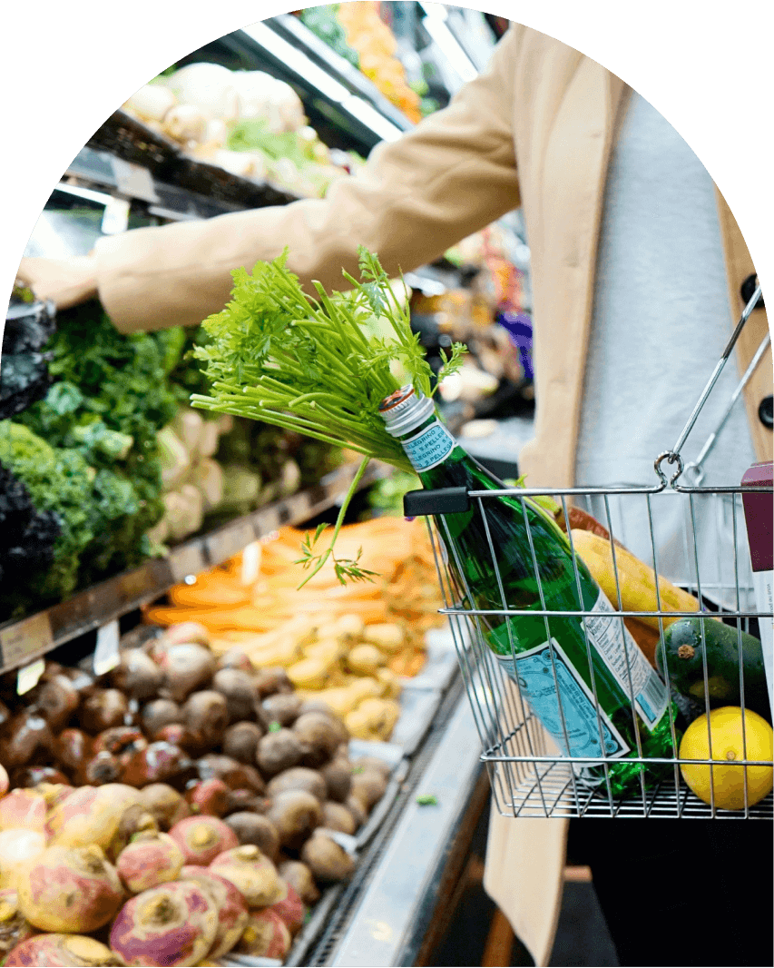 Person shopping for vegetables with a basket holding a water bottle, greens, cucumber, lemon, and bread.