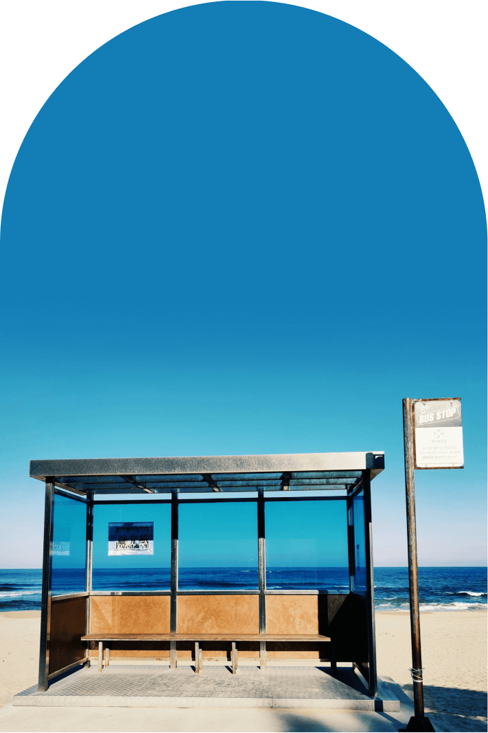 Empty bus stop shelter on sandy beach with blue ocean and clear sky in background.
