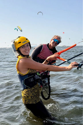 Two people smiling while kite surfing in the ocean, wearing helmets and harnesses.