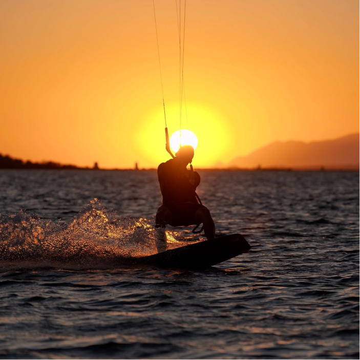 Silhouette of a person kiteboarding on water at sunset with sun near the horizon.