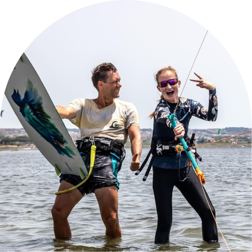 A man with a surfboard and a woman with a kiteboarding control bar stand smiling in shallow water.
