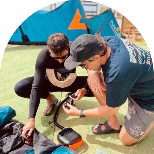 Two men kneeling on turf setting up kiteboarding equipment including a pump and kite valves.