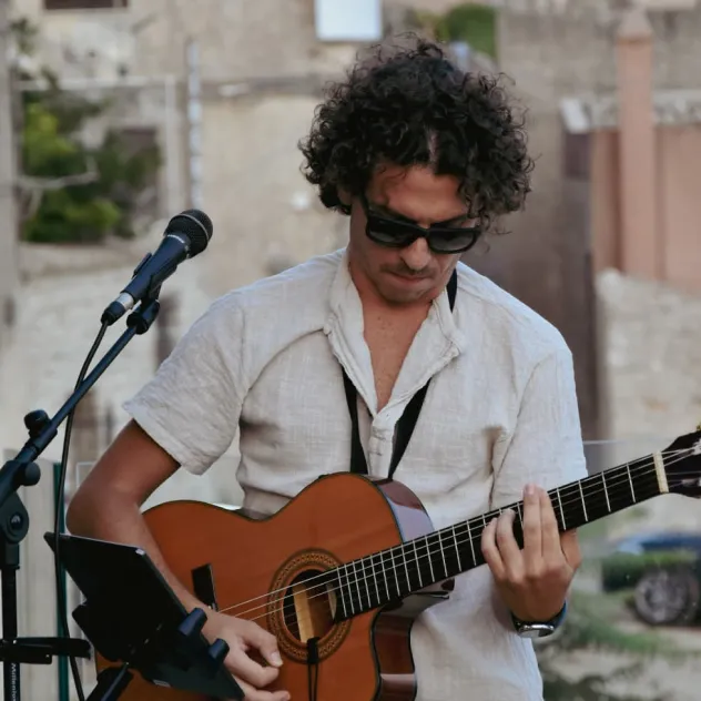 Man with curly hair and sunglasses playing acoustic guitar near a microphone outdoors.