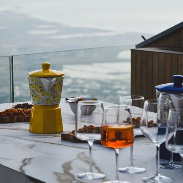 Table with a yellow coffee maker, glass of amber drink, and snacks with a blurred outdoor view.