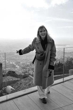 Woman smiling and leaning on a glass railing with a city view and landscape behind her.