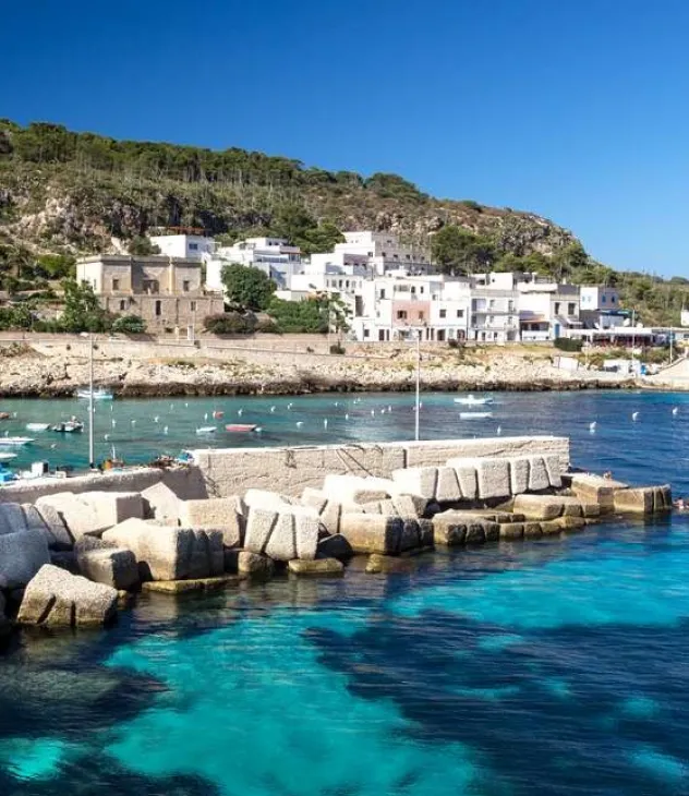 Clear blue water and stone pier with boats near a coastal village under sunny sky and green hillside.