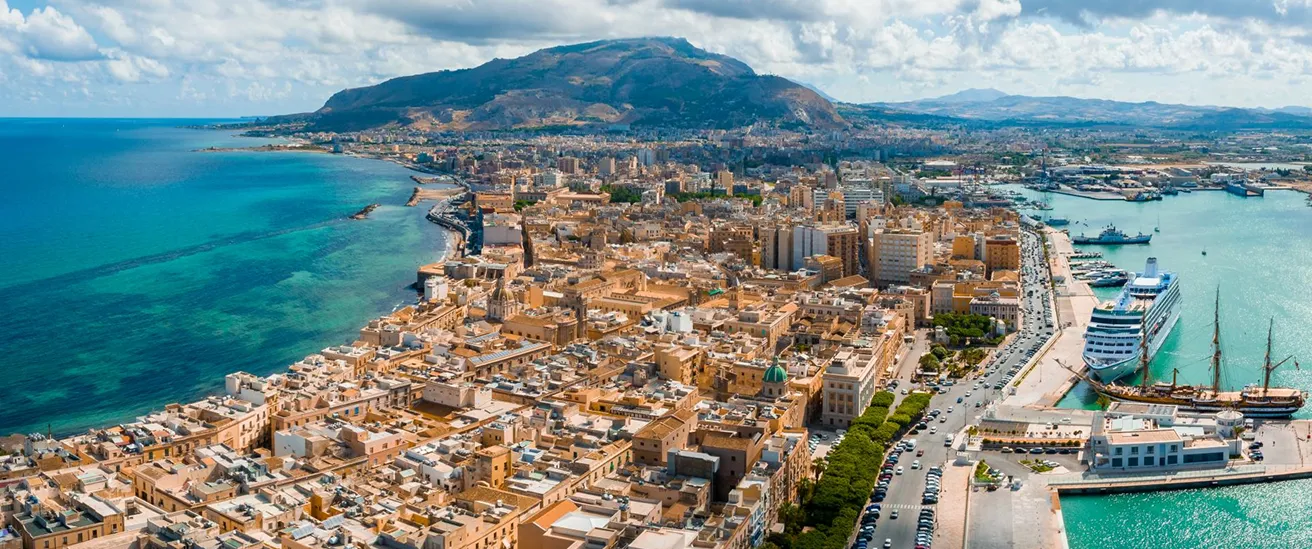 Aerial view of Trapani, Sicily, showing buildings, coastline, harbor with ships, and distant mountains.