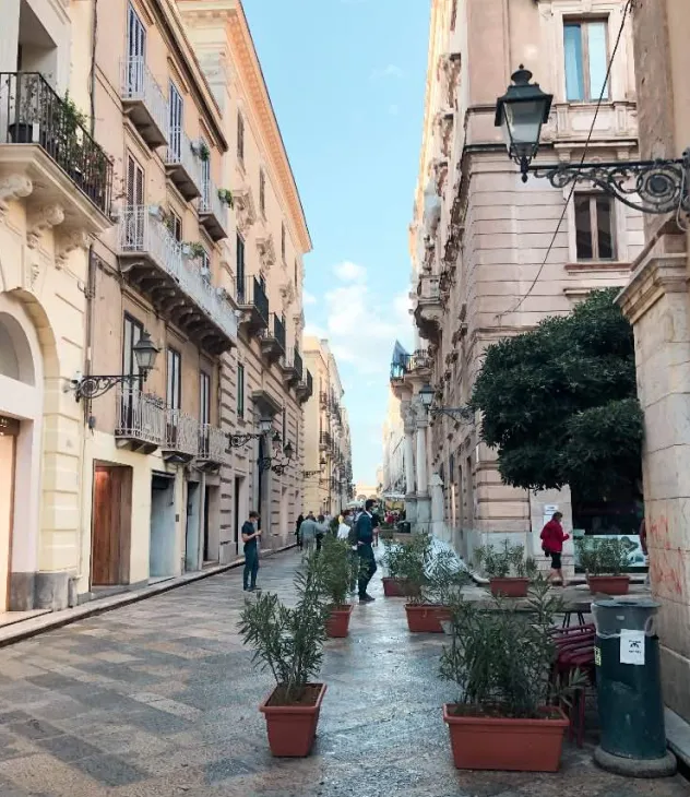 Narrow street with historic buildings, pedestrians, and potted plants under a blue sky.