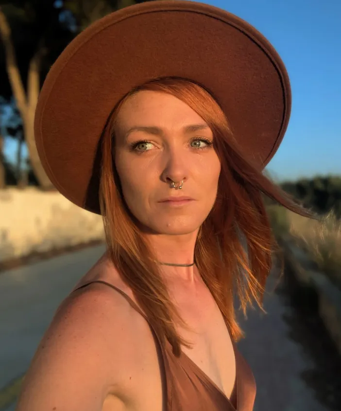 Woman with red hair wearing a brown hat and dress, standing outdoors in sunlight.