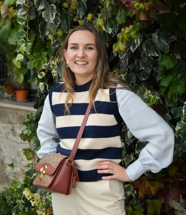 Smiling woman with striped sweater and beige pants standing in front of leafy green plants.