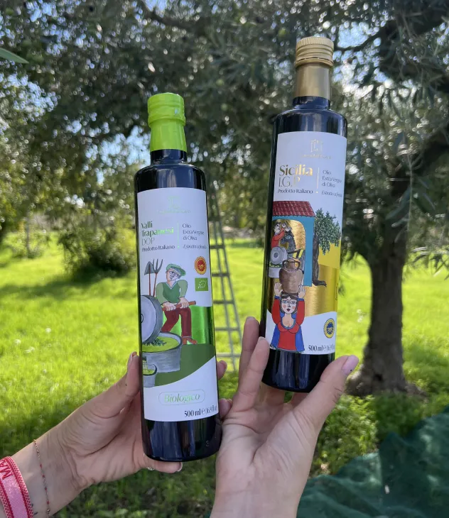 Two hands holding two bottles of Italian extra virgin olive oil outdoors with olive trees in background.