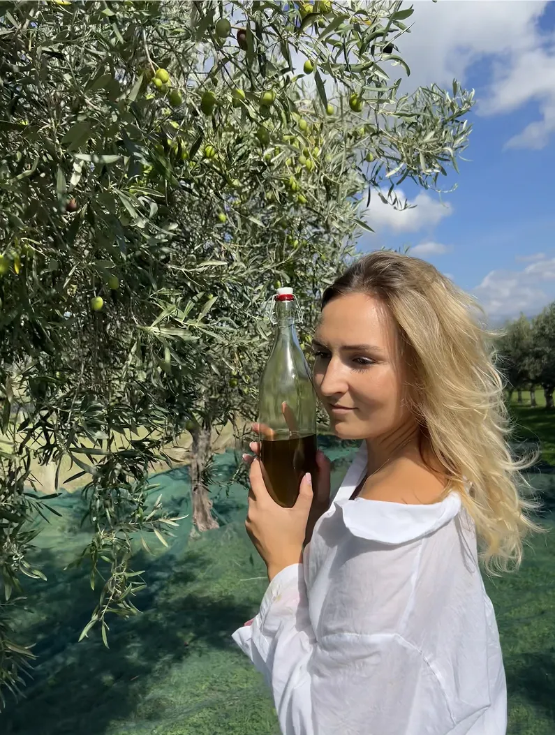 Woman holding a glass bottle of olive oil beside an olive tree in a sunlit orchard.