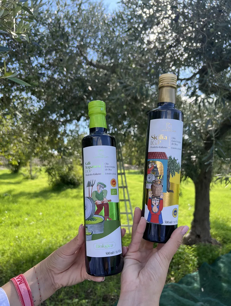 Two hands holding bottles of Italian extra virgin olive oil with orchard trees in the background.
