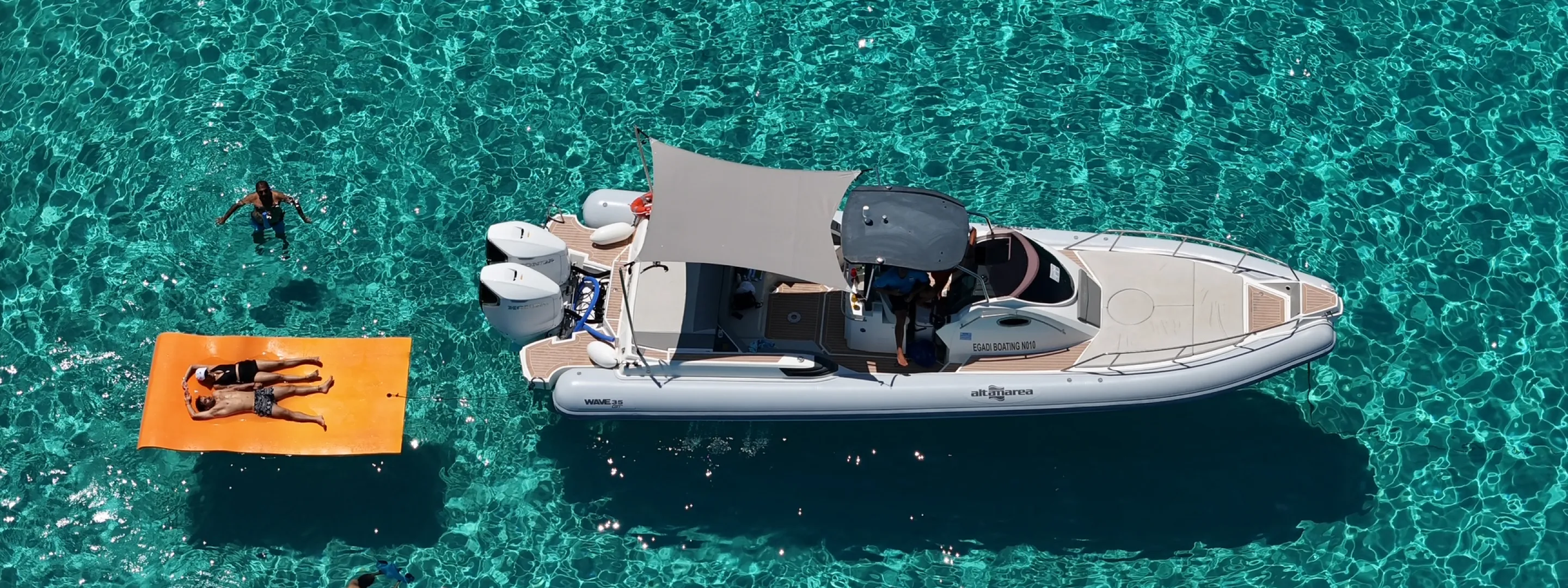 White motorboat with canopy floating on clear turquoise water with two people on an orange float nearby.