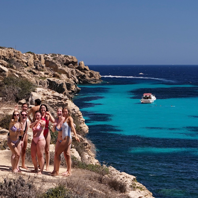 Group of people in swimsuits waving from rocky shore with turquoise sea and boat in background.