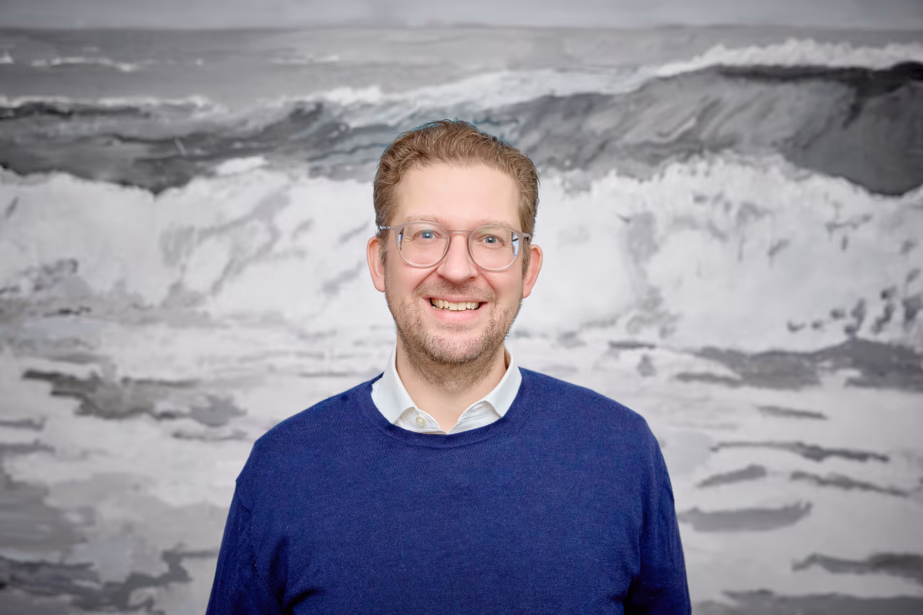 Lächelnder Mann mit Brille, weißem Hemd und blauem Pullover vor einem abstrakten schwarz-weißen Hintergrund.