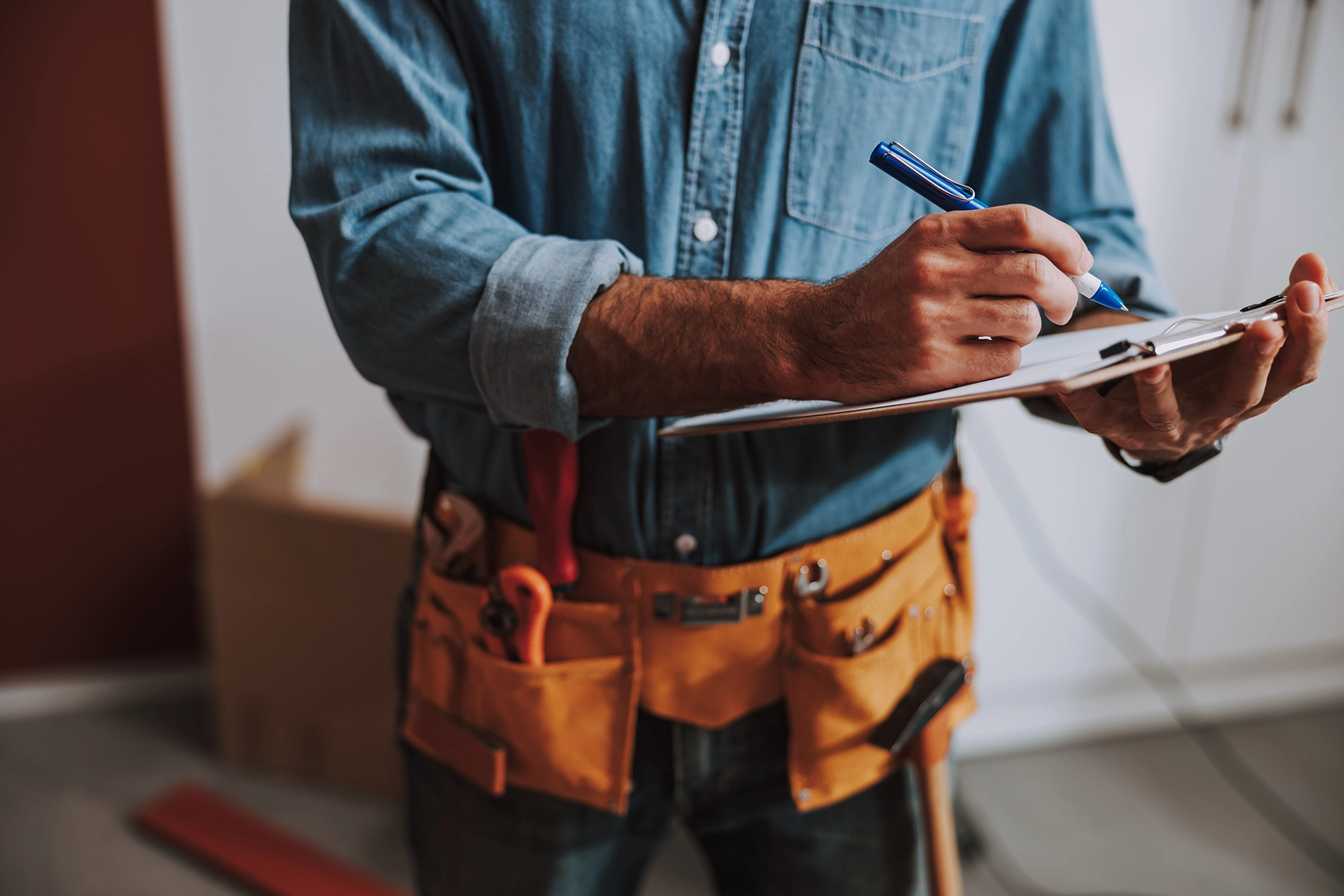 Person in blue denim shirt with a tool belt writing on a clipboard with a blue pen. Peak Pro Home Service Team Member.