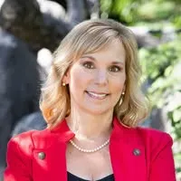 Smiling woman with blonde hair wearing a red blazer and pearl necklace outdoors with green foliage in the background.