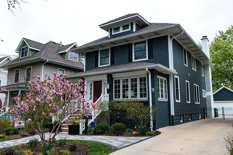 Two-story dark gray house with white trim, front porch with red door, and a flowering tree in the front yard. Typical Peak Pro Home Services client home.