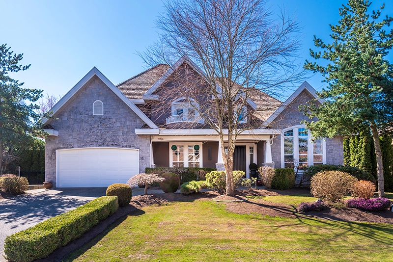 Large suburban house with stone exterior, two-car garage, and landscaped front yard with trees and bushes. Typical Peak Pro Home Services client home.