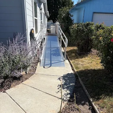 Aluminum wheelchair ramp with handrails installed alongside a house, connecting a sidewalk to the house entrance, surrounded by greenery and a lawn.