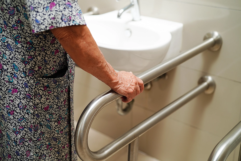 Senior person holding onto a stainless steel grab bar next to a bathroom sink.