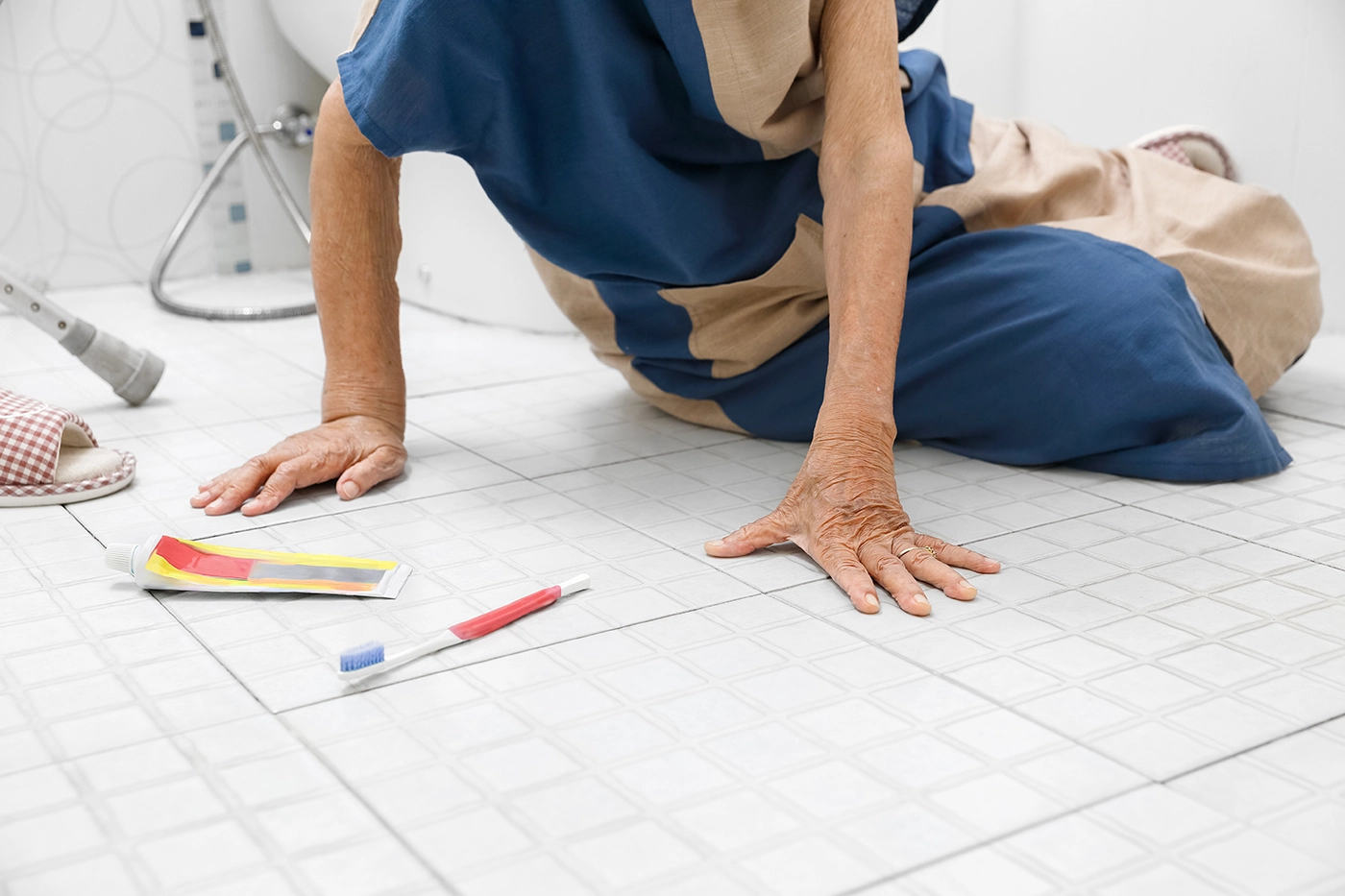 Senior person on the bathroom floor reaching toward a dropped toothbrush, illustrating a fall risk caused by slippery surfaces during daily routines.