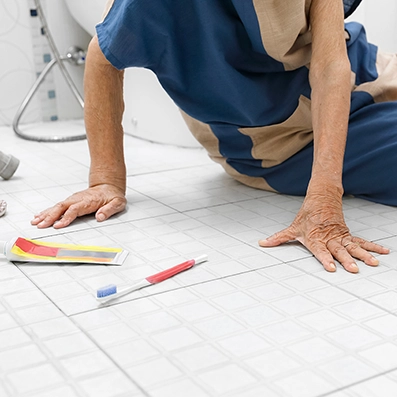 Senior person on the bathroom floor reaching toward a dropped toothbrush, illustrating a fall risk caused by slippery surfaces during daily routines.