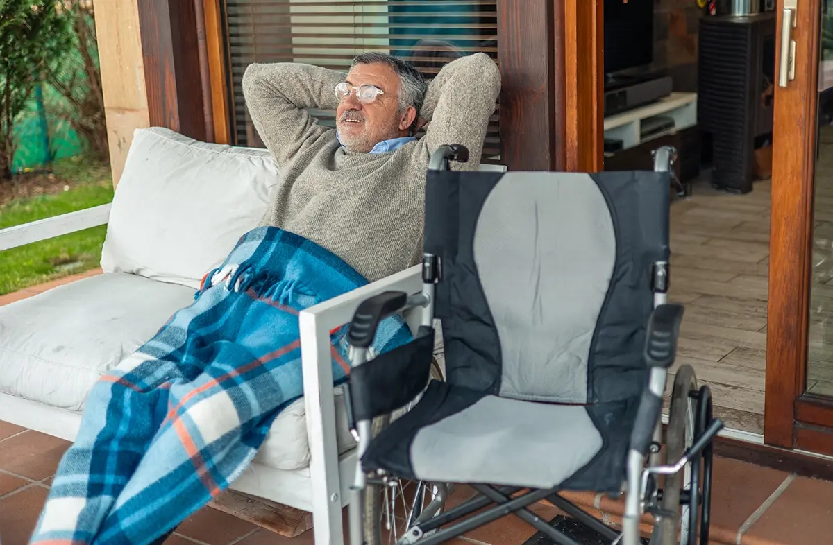 An older man relaxes on his covered home porch with a blanket over his legs and his wheelchair beside him.