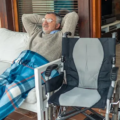 An older man relaxes on his covered home porch with a blanket over his legs and his wheelchair beside him.