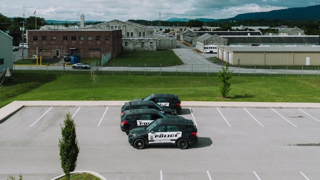 Three police SUVs parked in an empty parking lot near industrial buildings and grassy areas under a cloudy sky.