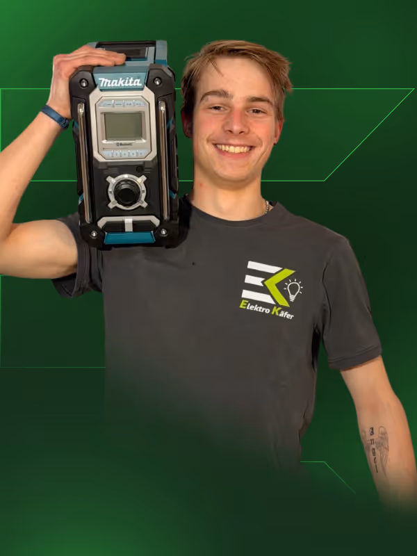 Junger Mann in grauem Elektro Käfer T-Shirt hält ein Makita Baustellenradio auf der Schulter und lächelt vor grünem Hintergrund.