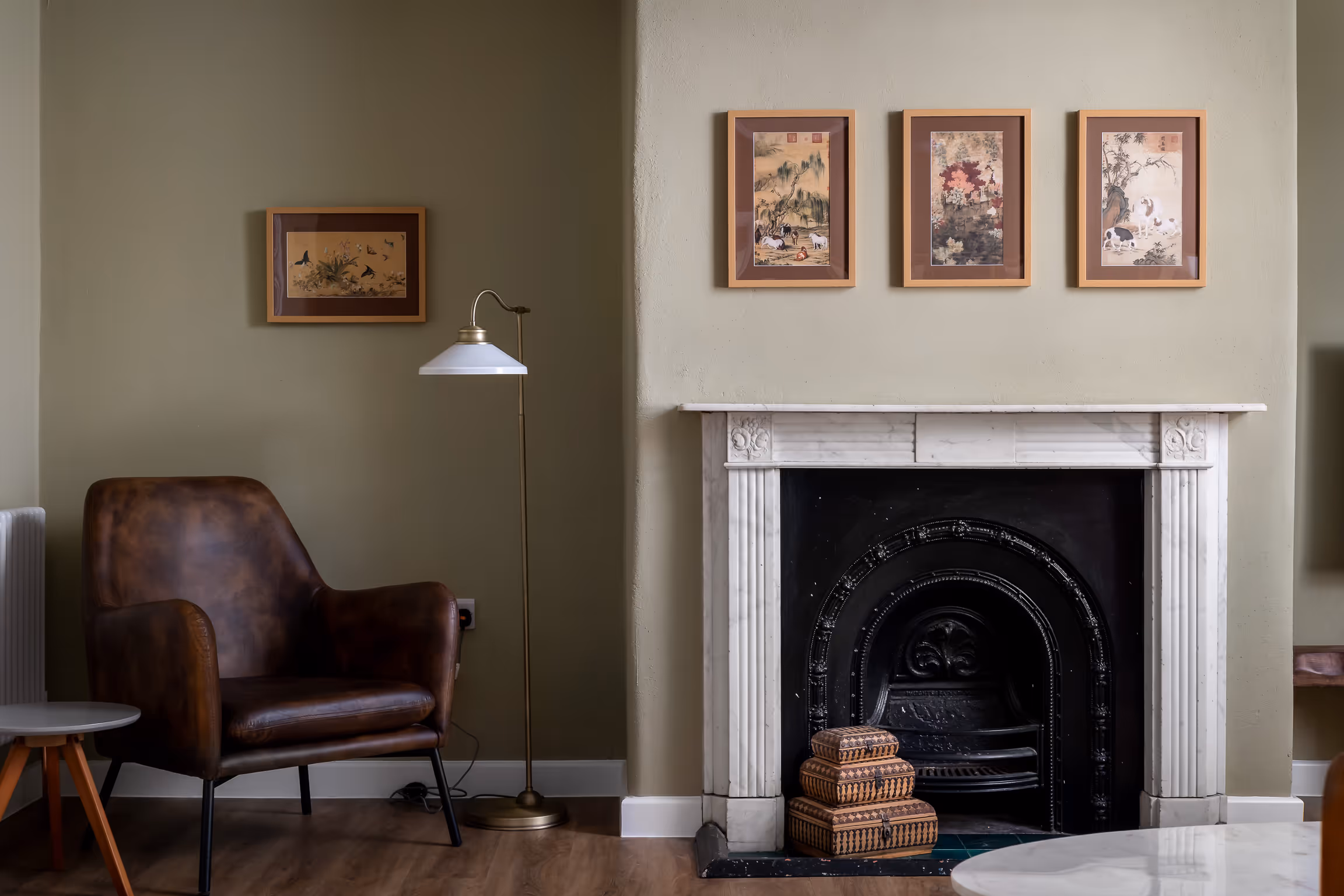 Cozy living room corner with a brown leather armchair, floor lamp, small round table, and a white marble fireplace with three framed artworks above.