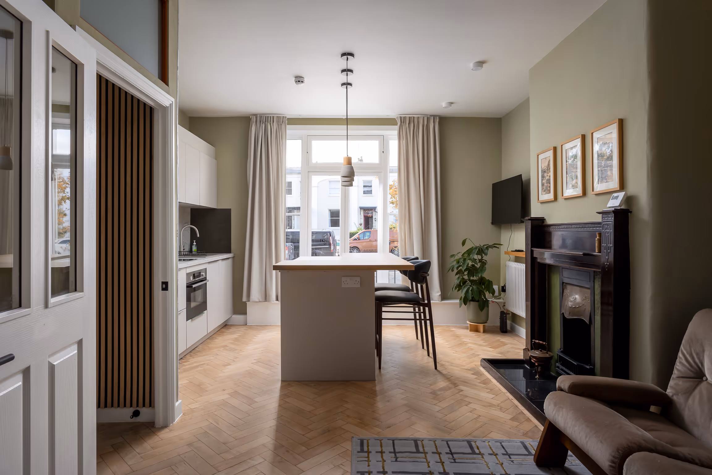 Modern kitchen and dining area with white cabinetry, an island with chairs, large window with curtains, green walls, fireplace, and beige armchair.