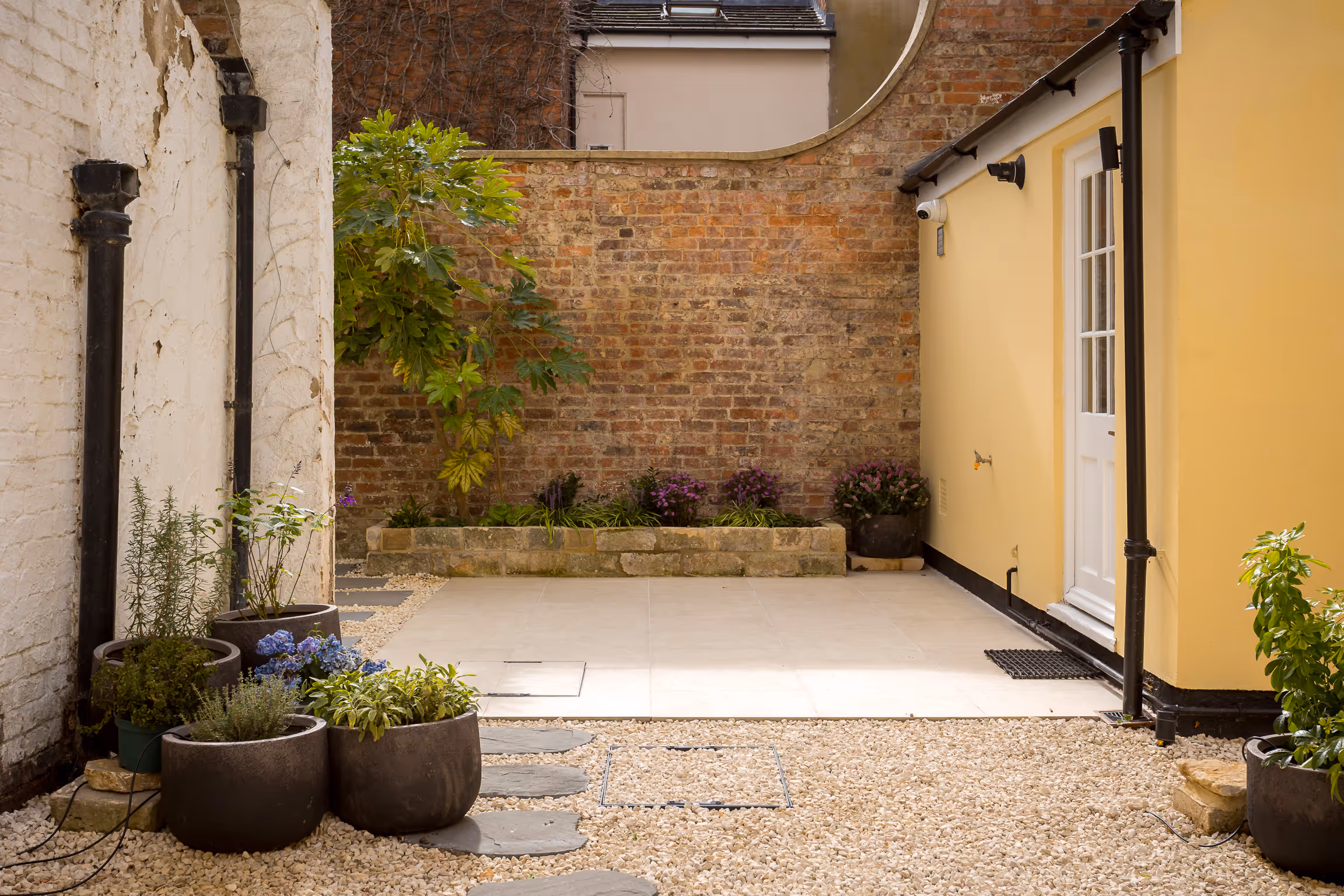 Small enclosed courtyard with gravel ground, stone stepping path, potted plants, a brick wall, and yellow building with a white door.