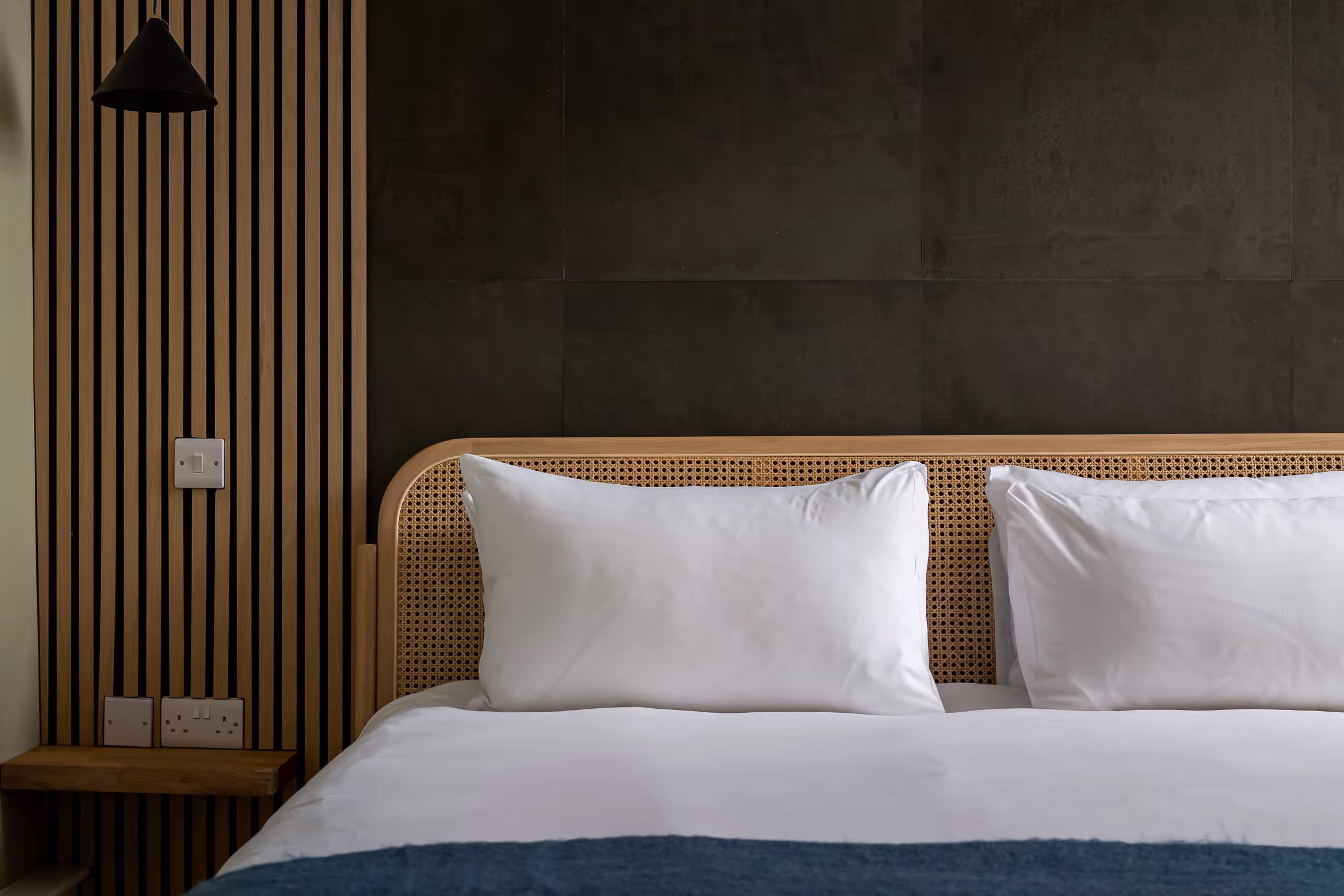 Modern bed with white pillows, white sheets, blue blanket, rattan headboard, and a vertical wooden slat wall with electrical outlets.