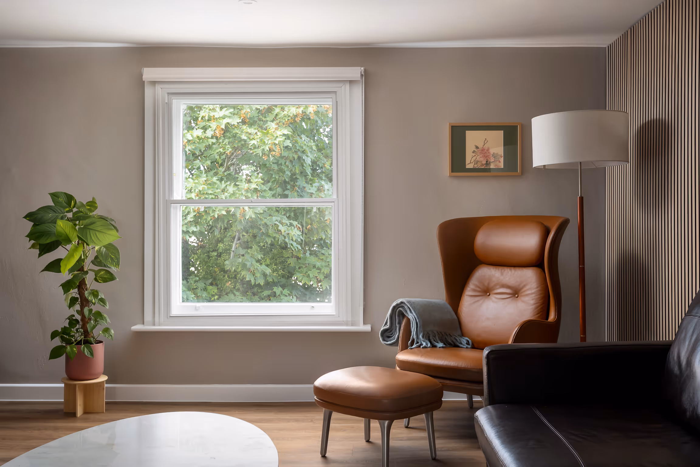 Cozy living room corner with a brown leather armchair, matching ottoman, floor lamp, potted plant, and large window showing green tree outside.
