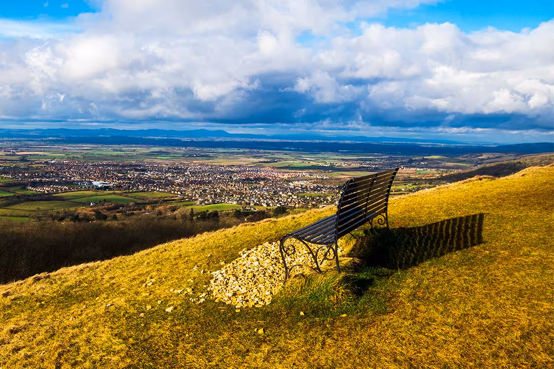 Empty metal bench on a grassy hill overlooking a town and distant fields under a partly cloudy sky.