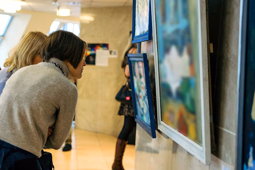 Two women closely examining framed paintings displayed on a gallery wall.
